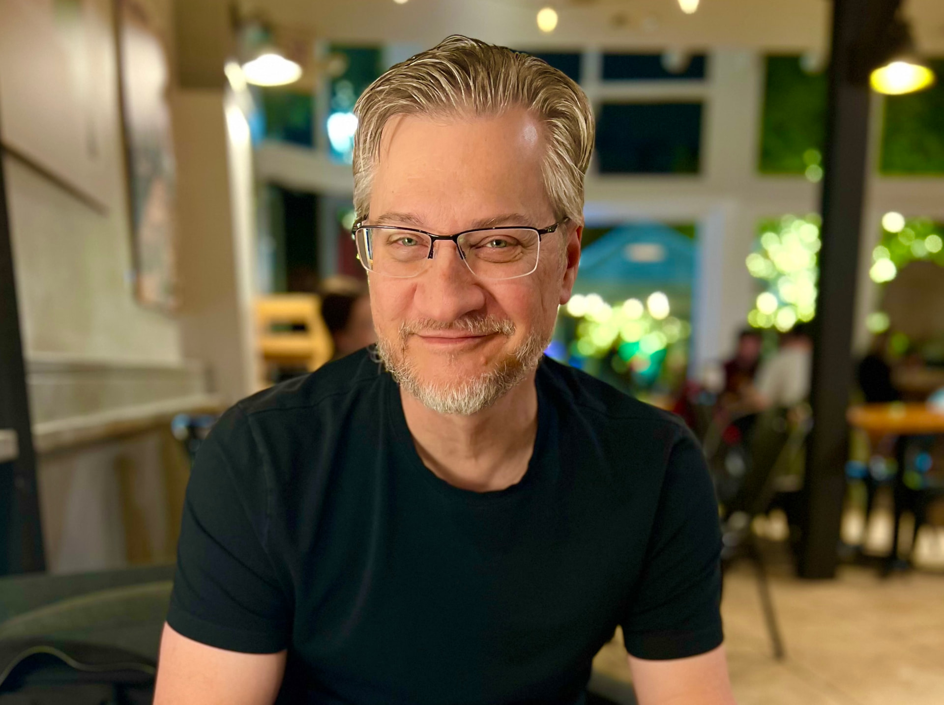 Tim Ristow smiling in a warm café, wearing glasses and a black shirt